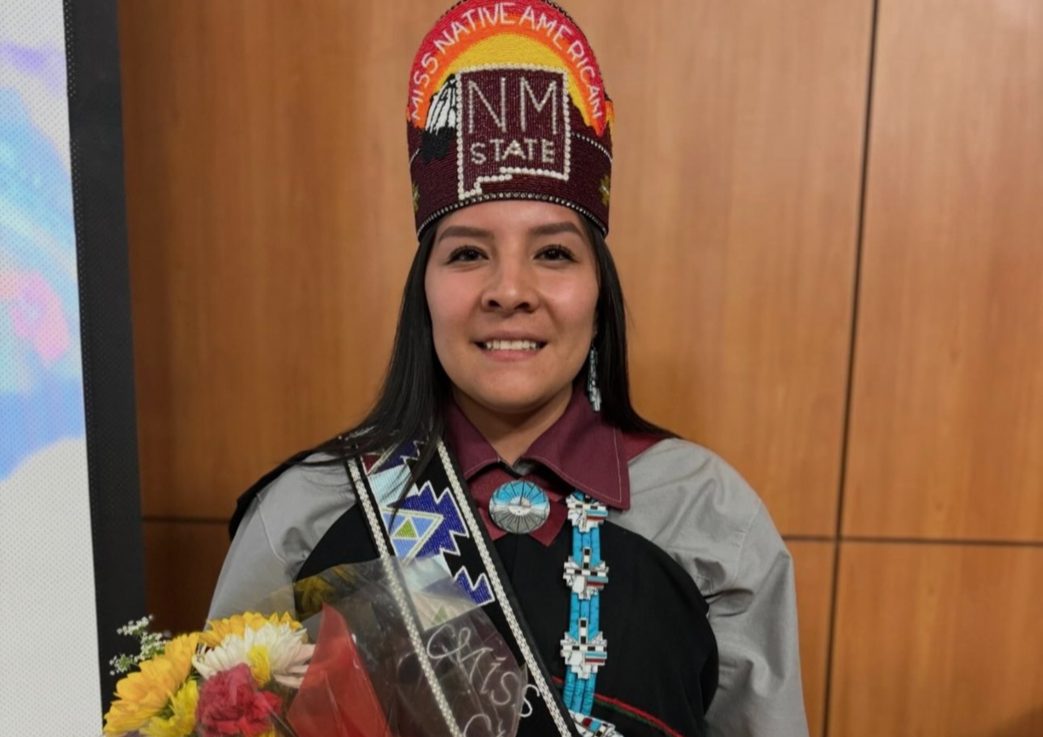 Ms Native American NMSU. Woman wearing traditional regalia, a crown with "MISS NATIVE AMERICAN" and "NM STATE," and holding a bouquet.